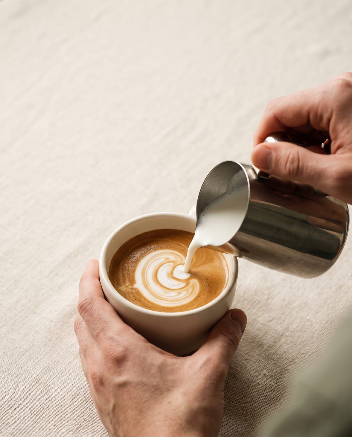 Tangan barista menuang susu ke dalam cup keramik putih.