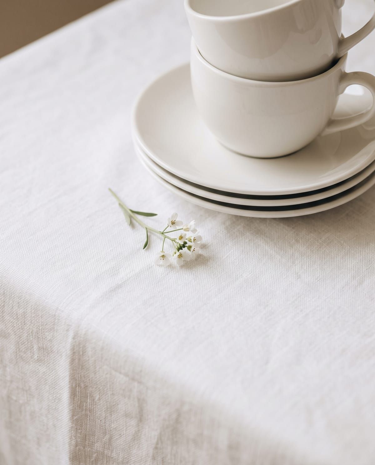 Tumpukan cup dan lepek keramik putih di atas taplak linen putih dengan setangkai bunga kecil.