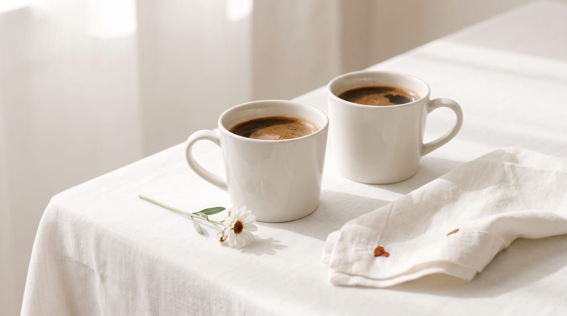 Dua cup kopi di atas taplak linen putih dengan setangkai bunga kecil.